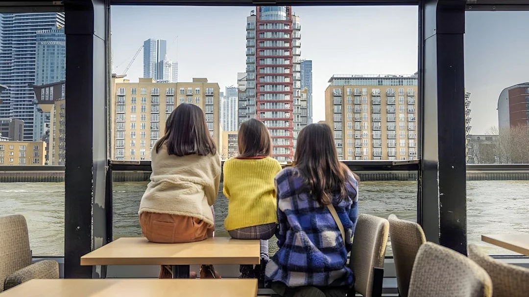 Three people seated inside a Thames River cruise boat, viewing modern London buildings.