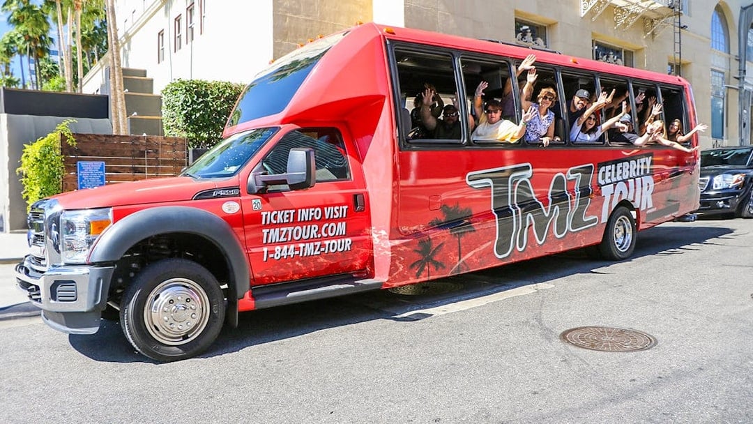 TMZ tour bus with passengers waving on Hollywood street.