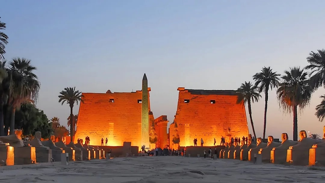 Luxor Temple entrance at dusk with illuminated obelisk and palm trees, Egypt.