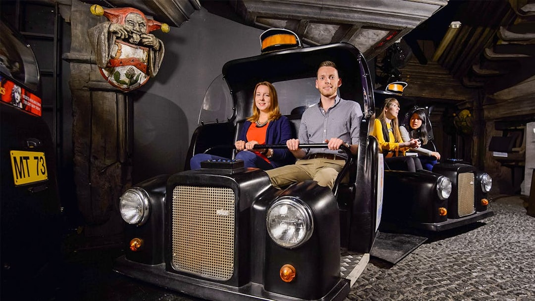 Visitors on Spirit of London Ride in black taxi-themed cars at an indoor attraction.