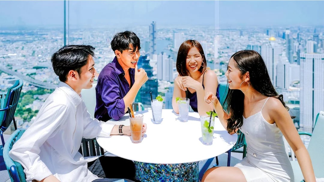 Friends enjoying drinks at Mahanakhon SkyWalk, Bangkok, with city skyline view.