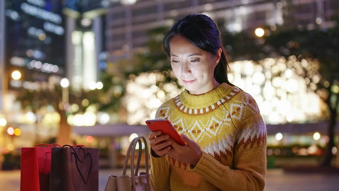 Woman using a smartphone in a city at night with shopping bags nearby.