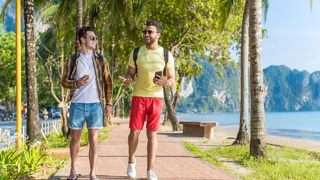 Two people walking on a palm-lined path by the beach, using phones in Kuala Lumpur, Malaysia.