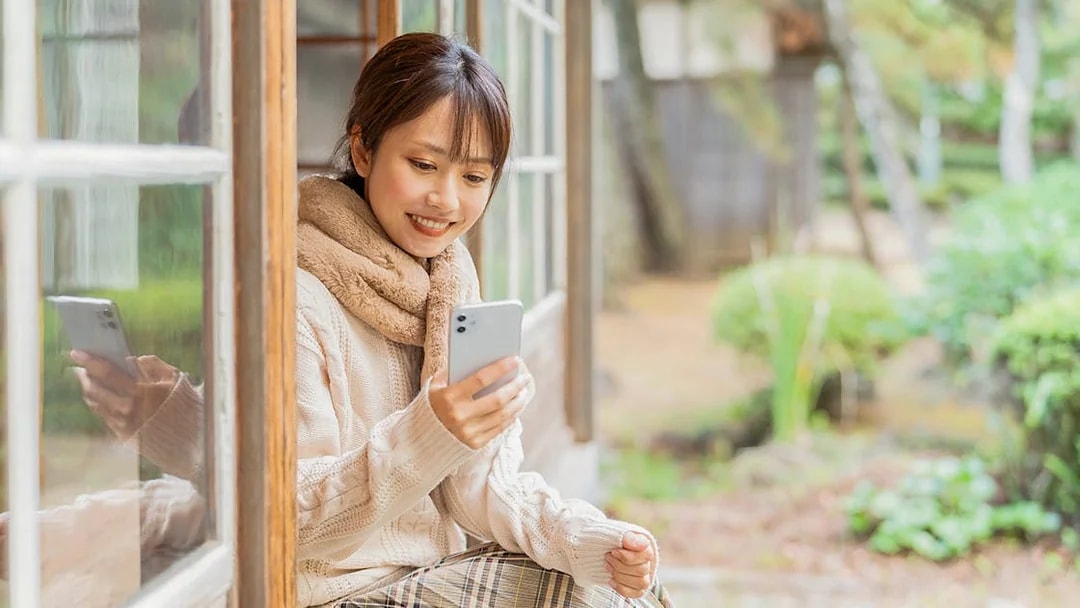 Woman sitting by a window, smiling while talking on a cellphone in a garden setting.