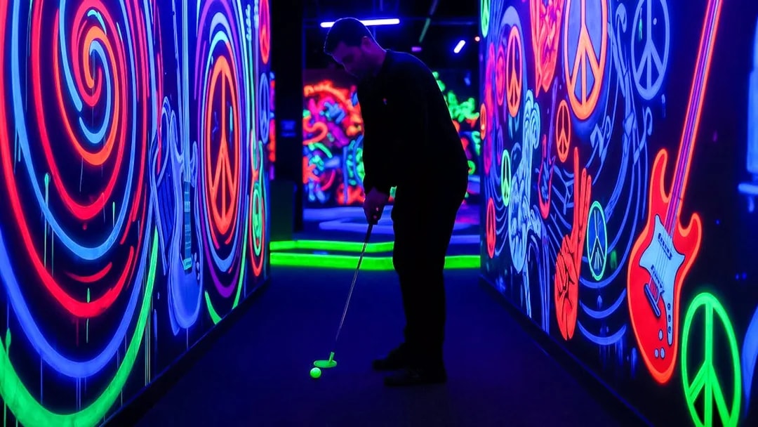 Man playing minigolf in a neon-lit Rock of Ages blacklight course with vibrant wall art.