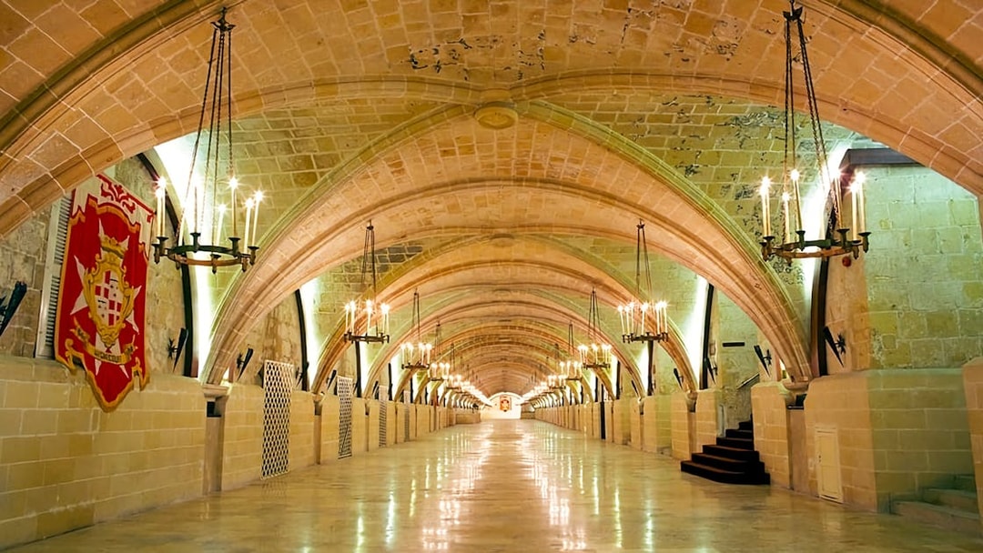 Grand arched hallway in the Holy Infirmary, Malta, with chandeliers and a Maltese cross banner.