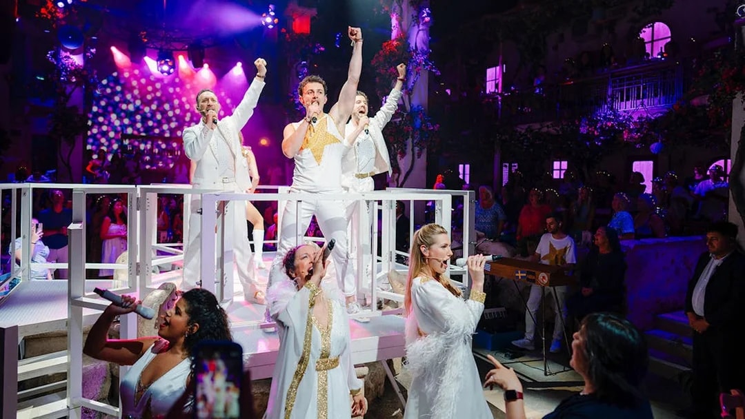 Performers singing on stage at Mamma Mia! The Party with vibrant lighting and audience in the background.