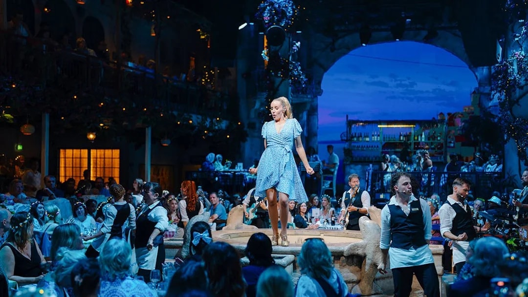 Performer on stage at Mamma Mia! The Party with audience dining in a Greek-themed setting.