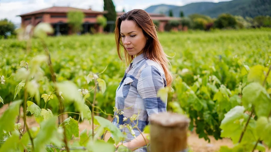 Young woman walking through a lush vineyard with a rustic building in the background.
