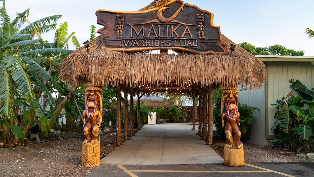 Entrance arch with dried grass at Mauka Warriors Luau, featuring carved wooden statues.