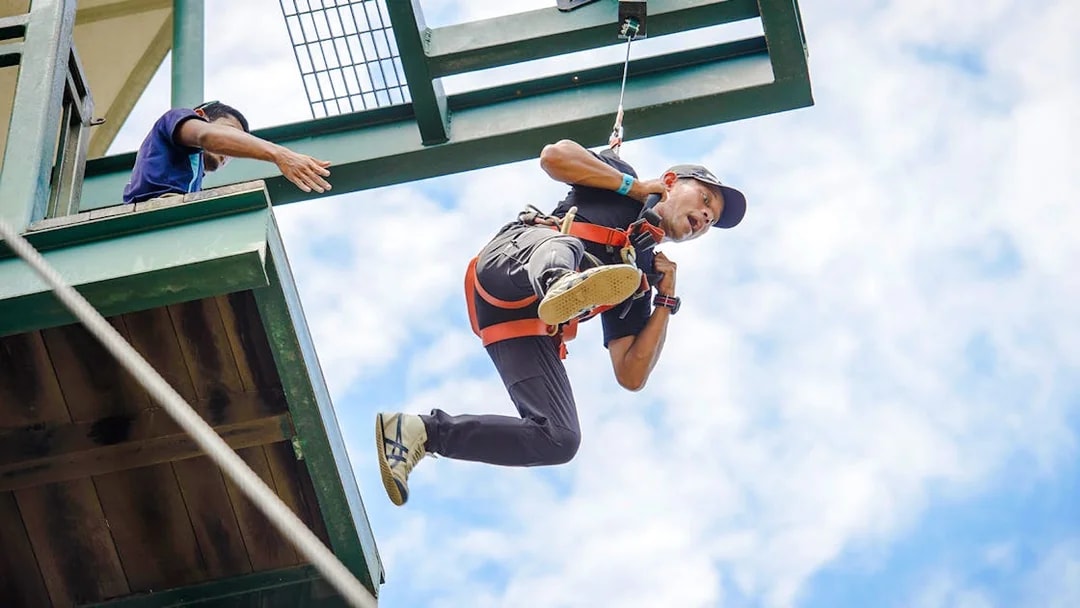 Person ziplining at Mega Adventure Park, Singapore against a blue sky.