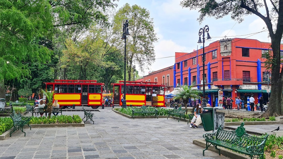 Red and yellow trams in Coyoacán square, Mexico City, with vibrant buildings and trees.
