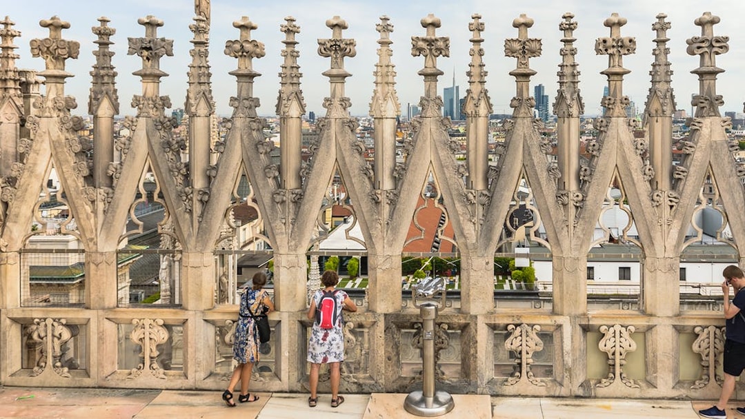 Visitors on the terraces of Milan Cathedral with city view through ornate stonework.