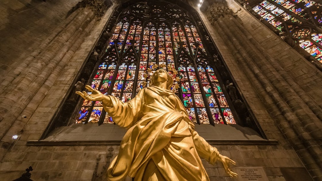 Golden statue inside Milan Duomo with stained glass windows in the background.