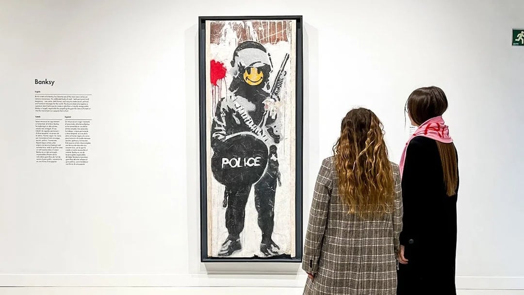 Tourists viewing Banksy artwork of a police figure with a smiley face.