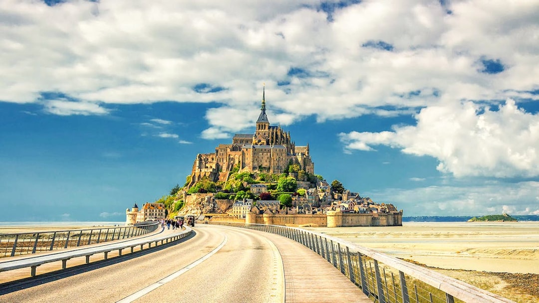 Mont Saint-Michel with bridge and visitors, France, under a blue sky.