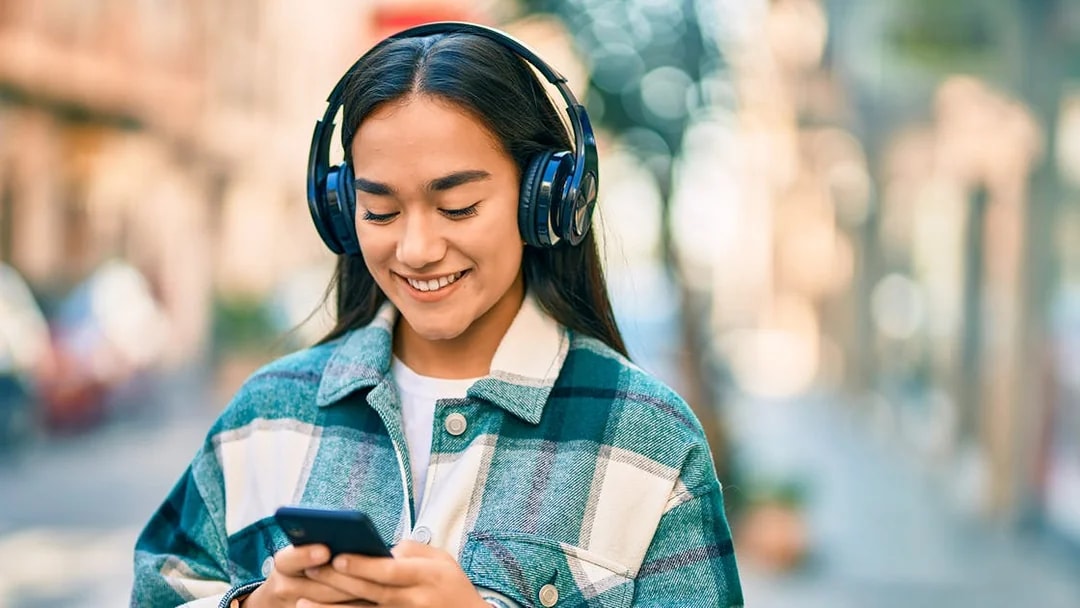 I don't know who this is, but here's the alt text:

"Person listening to music on headphones while using a smartphone outdoors.