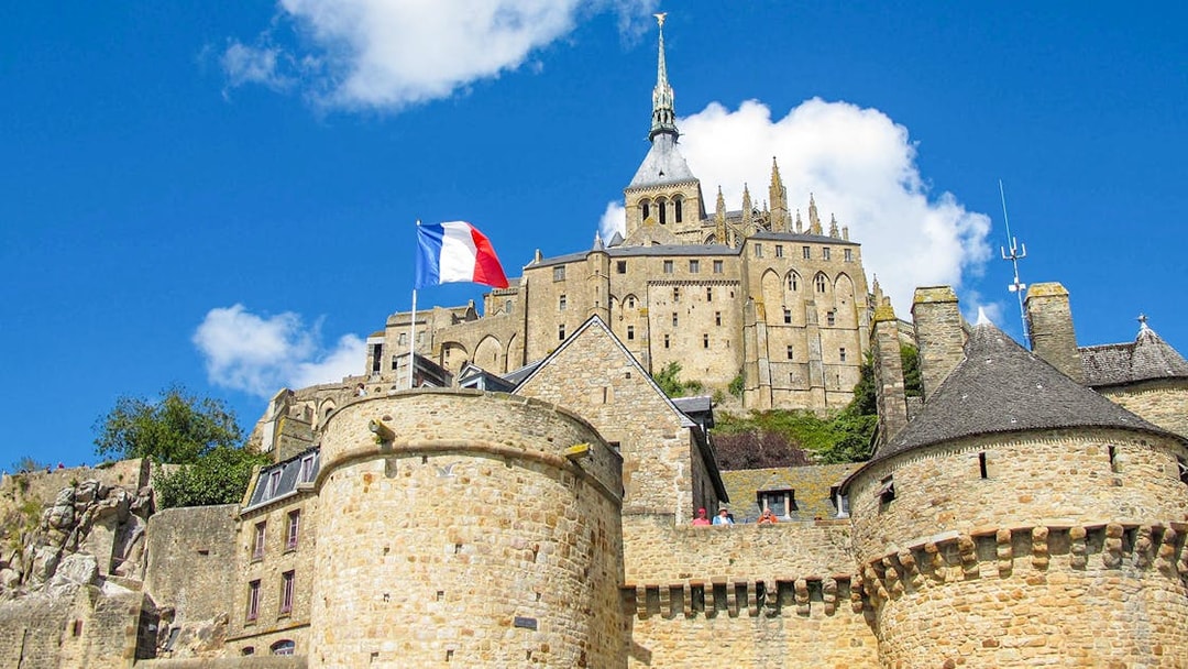 Mont Saint-Michel with French flag, stone walls, and abbey spire under blue sky.