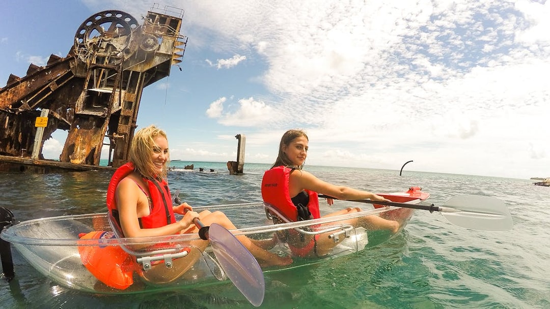 Two women kayaking in transparent kayaks near a rusted shipwreck in clear ocean waters.