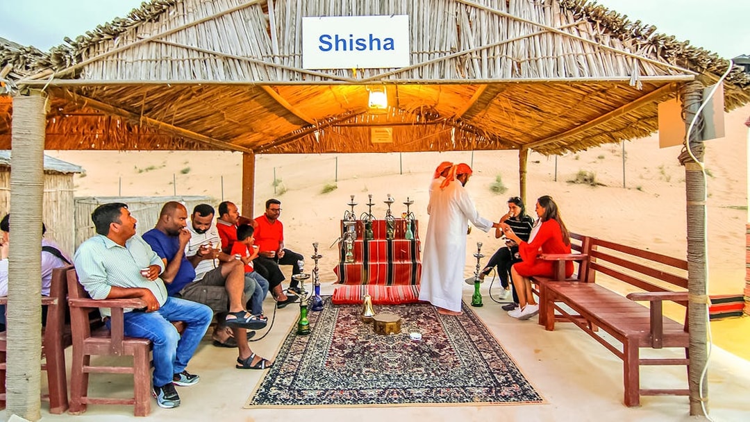 Desert safari guests enjoying shisha under a thatched roof in a sandy setting.