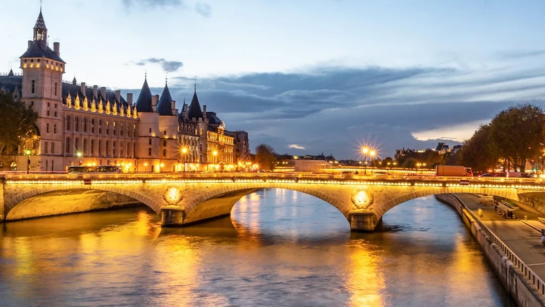 Conciergerie Paris illuminated at night with Seine River and bridge in view.
