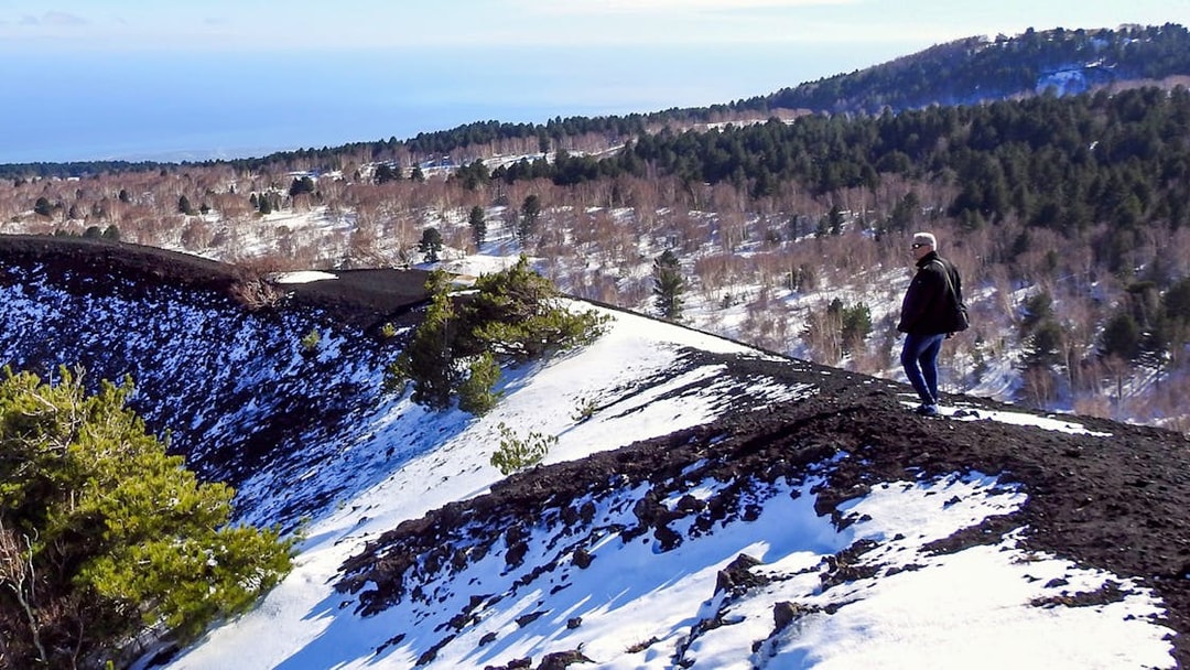 Person walking on snowy slope of Mount Etna with forested landscape in background.