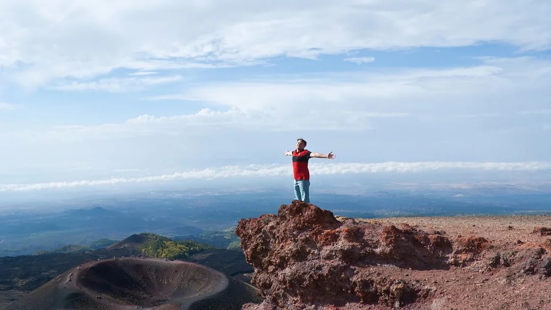 Person standing on rocky edge of Mount Etna with arms outstretched, Catania, Italy.