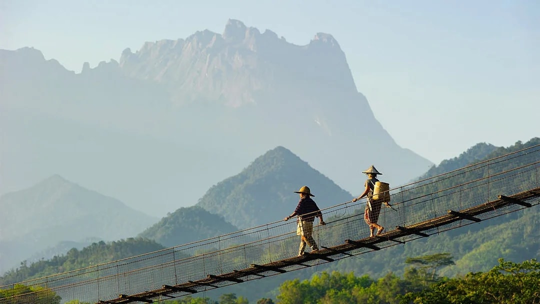 Hikers crossing a suspension bridge with Mount Kinabalu in the background.