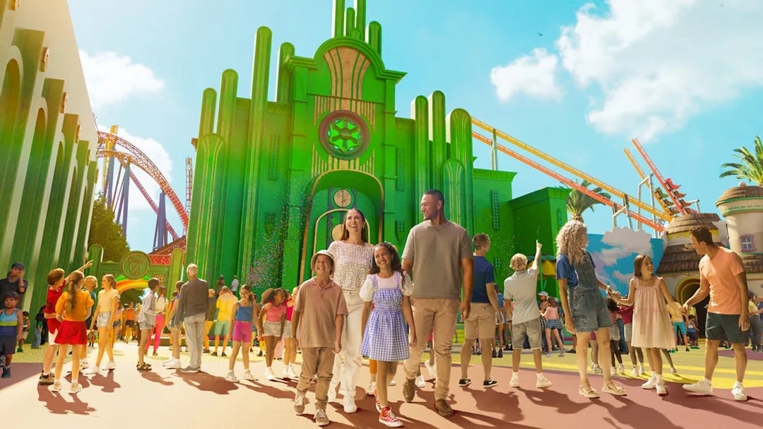 Tourists exploring Wizard of Oz precinct at Movie World with green buildings and roller coaster.