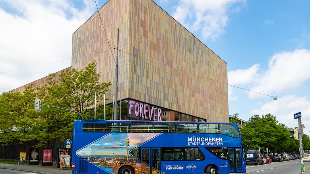 Blue double-decker tour bus in Munich near a modern art museum, part of city and FC Bayern Munich arena tour.