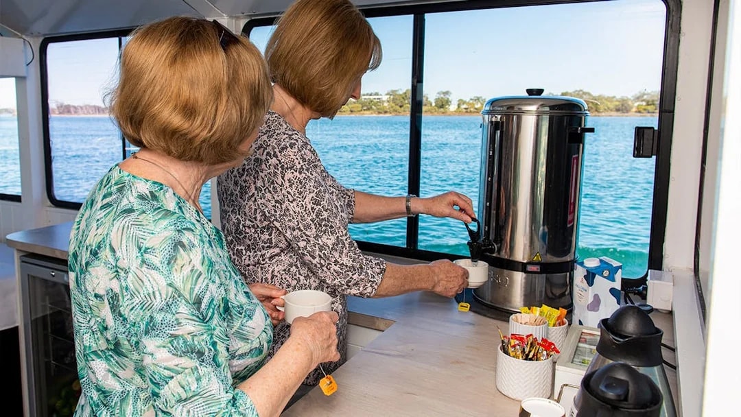Two people preparing tea on a Murray River lunch cruise with a scenic water view.