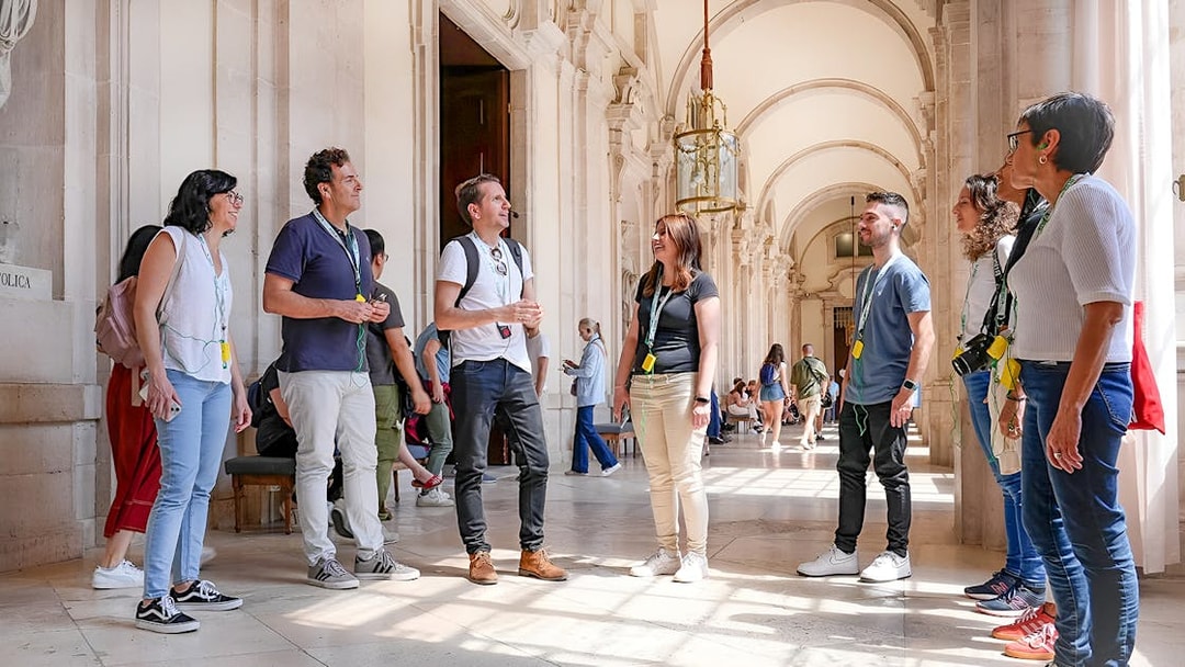 Group on a guided tour at the Prado Museum, Madrid, listening to a guide in a sunlit hallway.
