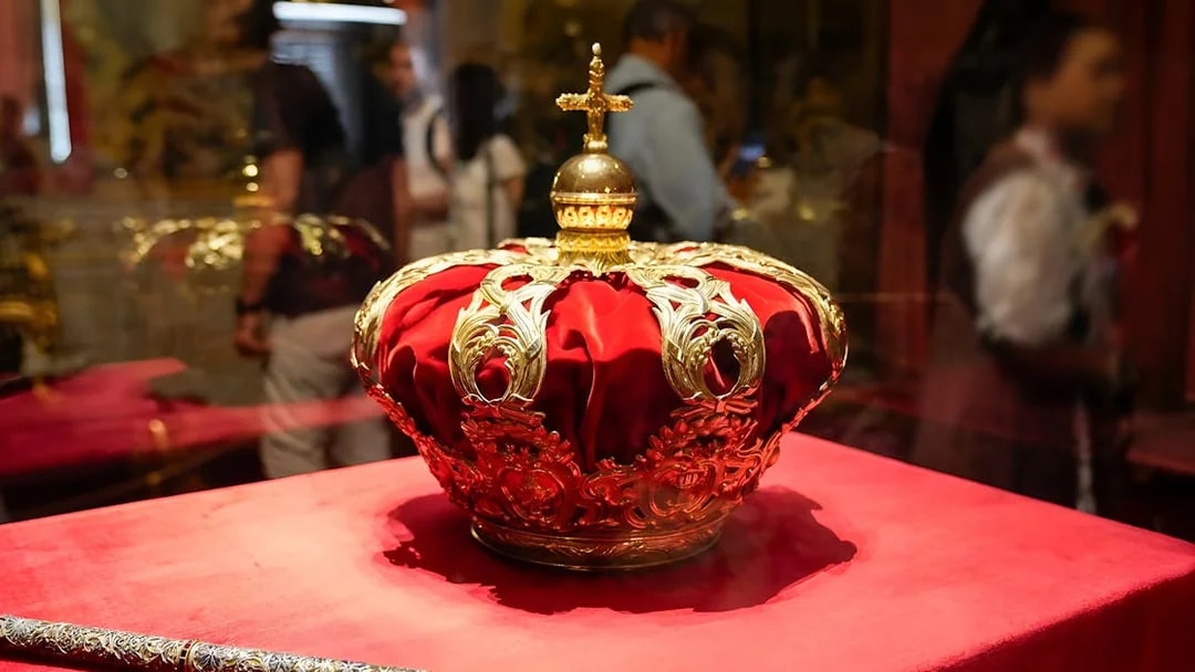 Royal crown on display at the Royal Palace of Madrid with tour group in background.