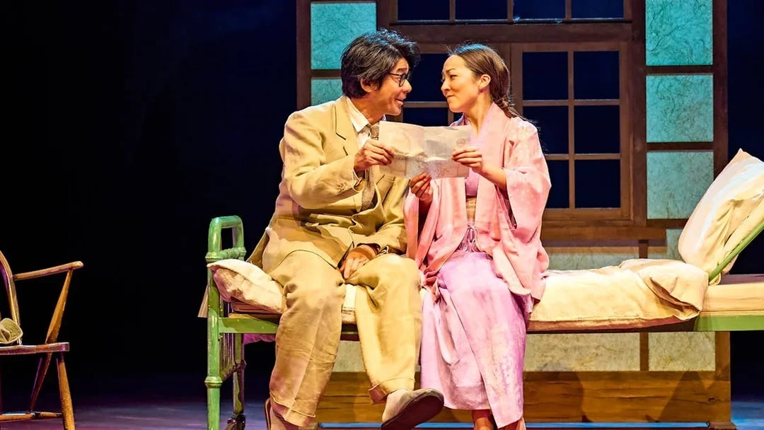 Two actors in a stage scene from "My Neighbour Totoro," sharing a moment on a bed.
