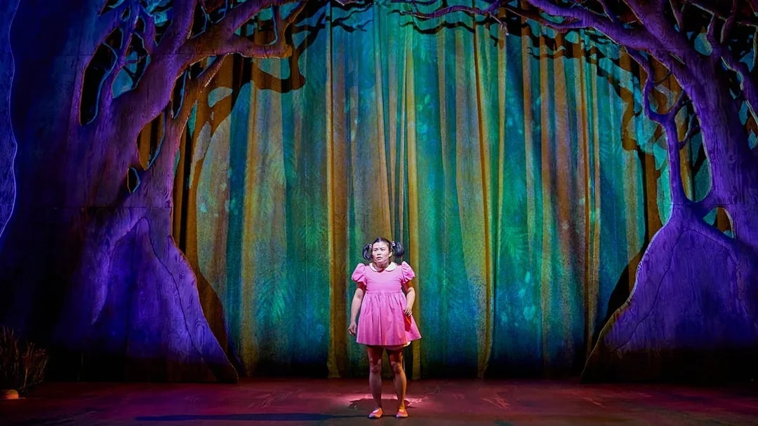 Stage scene from "My Neighbour Totoro" with character in pink dress under tree backdrop.