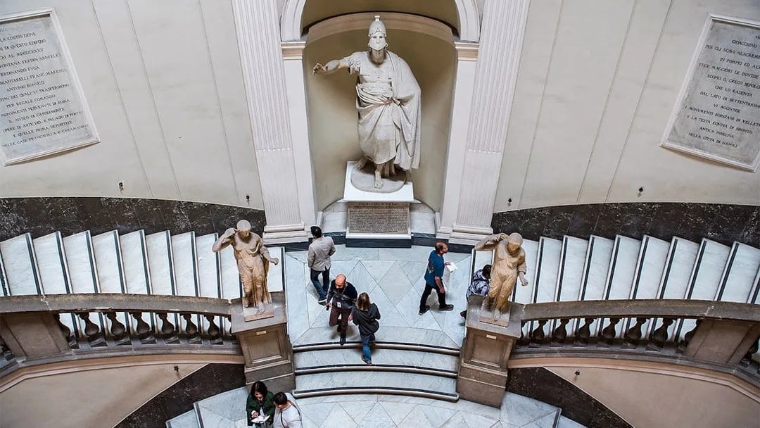 Visitors exploring statues and grand staircase at Archaeological Museum of Naples.