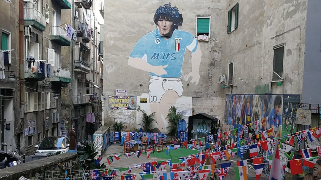 Mural of Diego Maradona in a Naples alley with international flags and memorabilia.