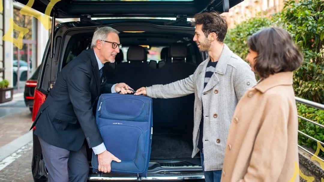 Chauffeur assisting couple with luggage into private airport transfer taxi.