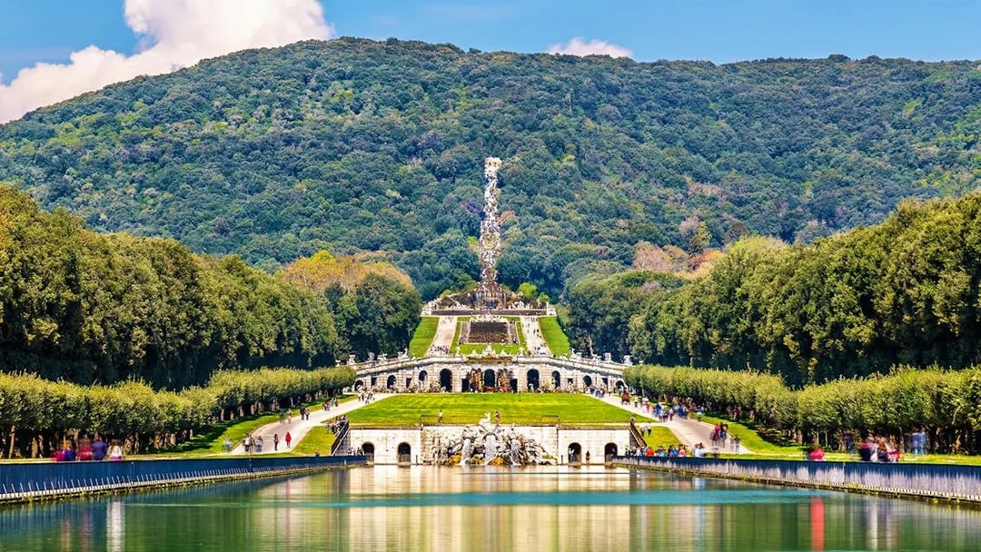 Royal Palace of Caserta gardens with fountain and wooded hills in the background.