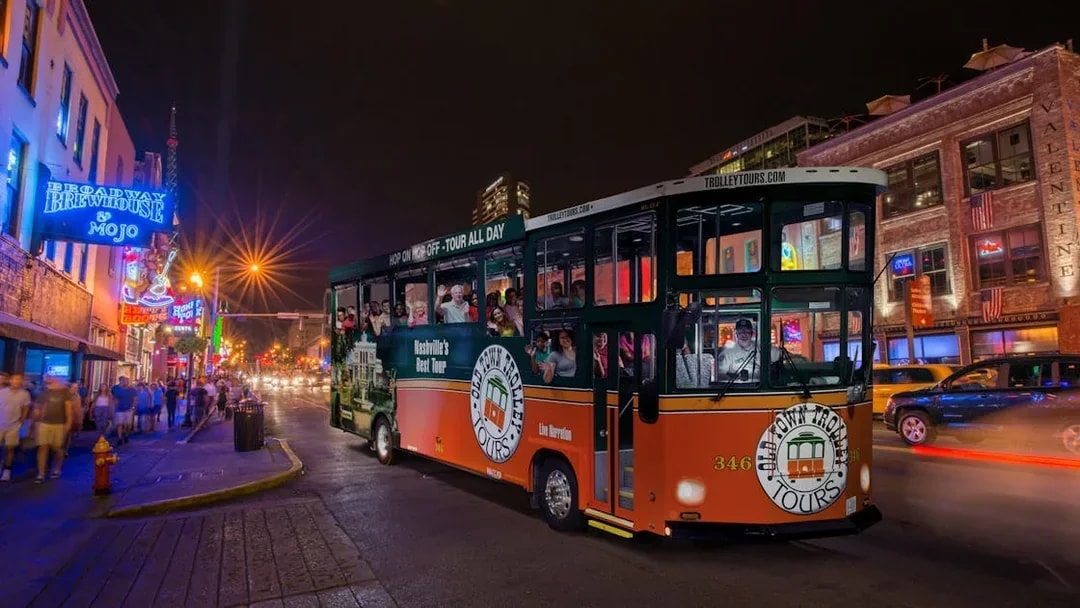 Hop-on hop-off trolley on a lively Nashville street at night, passing neon-lit bars and restaurants.