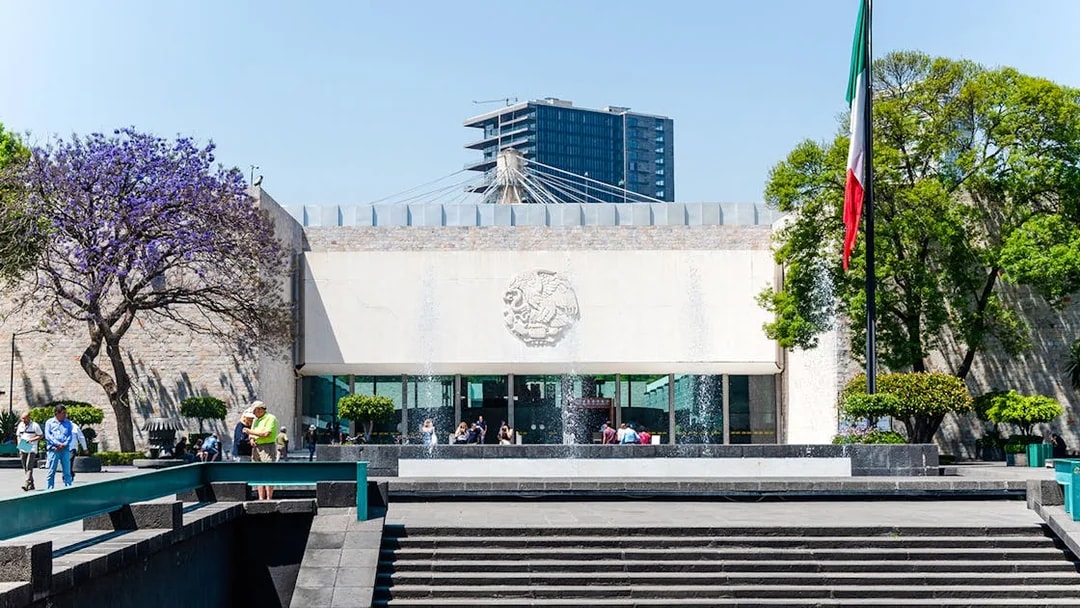 Main entrance of the Anthropology Museum in Mexico City with visitors and Mexican flag.