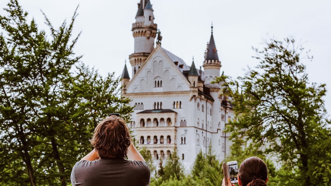 Visitors photographing Neuschwanstein Castle through trees in Bavaria, Germany.