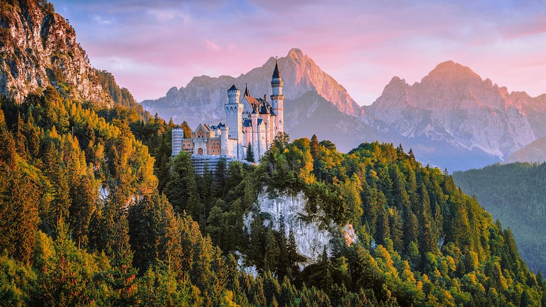 Neuschwanstein Castle aerial view at sunset, surrounded by forested mountains.