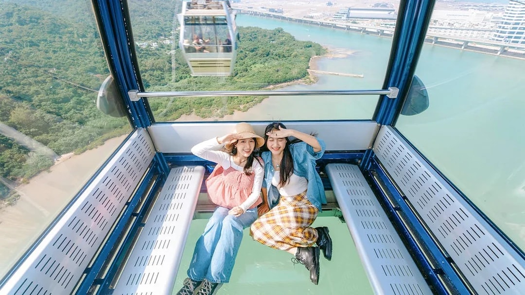 Two people sitting inside a crystal cabin cable car with a view of lush greenery and water below.