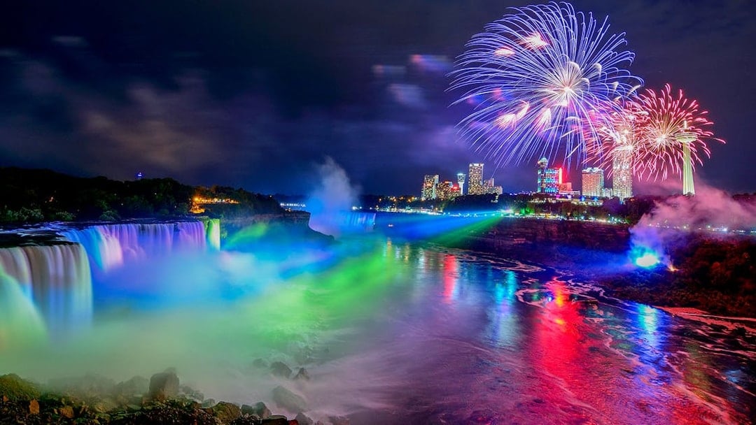 Niagara Falls illuminated at night with fireworks and city skyline in the background.