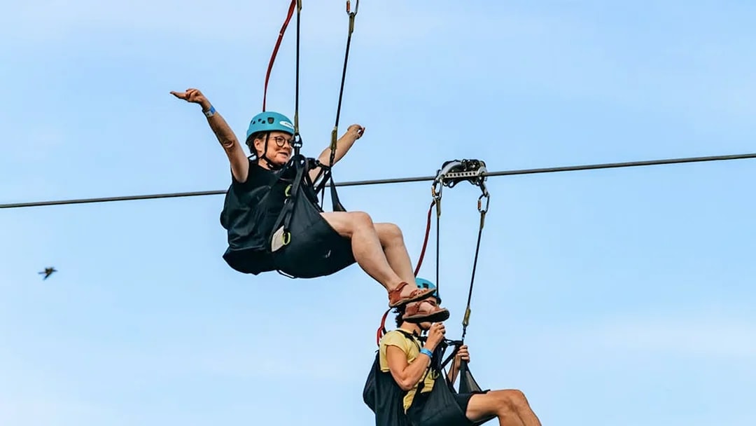 Two people ziplining with helmets and harnesses against a clear sky, part of the Zipline To The Falls experience.