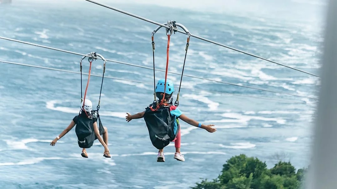 Two people ziplining over water towards a waterfall, wearing helmets and harnesses.