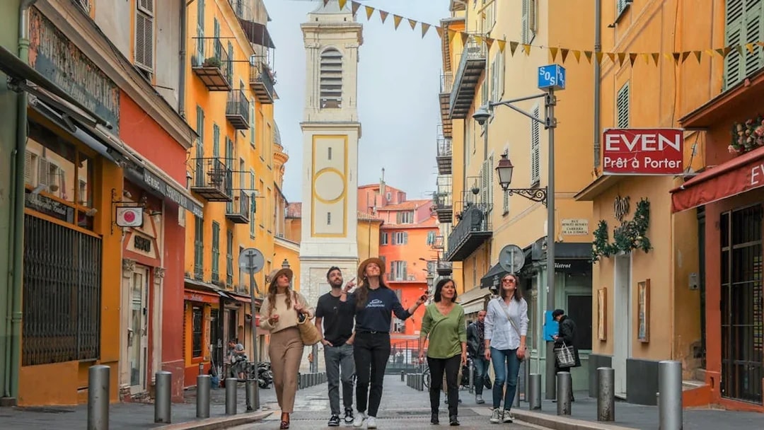 Group walking through colorful street in Nice with bell tower in background.