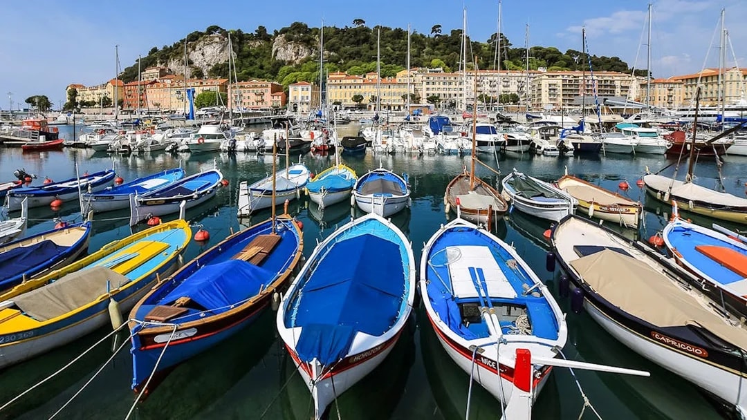 Colorful boats docked at Nice harbor with hillside buildings in the background.