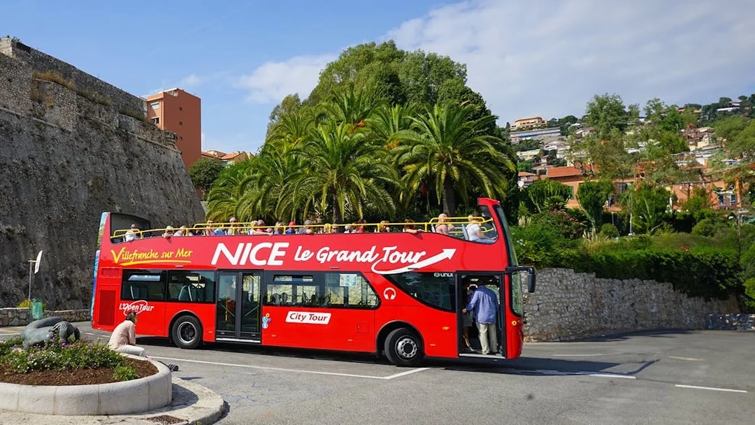 Red double-decker bus on Nice & Villefranche hop-on hop-off tour with palm trees in background.
